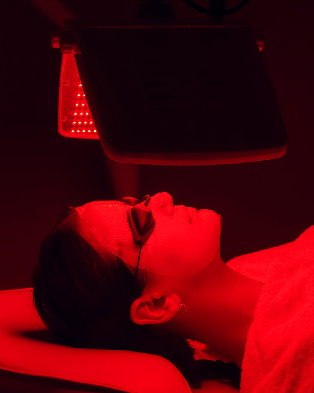 A lady laying on a bautician's bed wearing a sheet masque and goggles, under an LED red light therapy.